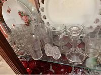 Photo showing a collection of etched glass stemware including glasses, goblets, floral decorative plates, glass candle holders, and a decanter stopper.
