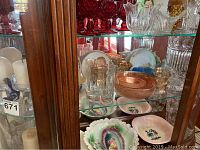 Display of orange carnival glass bowls, glasses, decorative plates, and clear ribbed vase on glass shelf