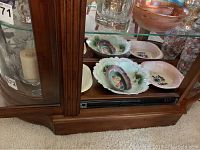 Wooden display cabinet bottom shelf with multiple vintage painted dishes, scalloped edges, floral designs in various colors and shapes