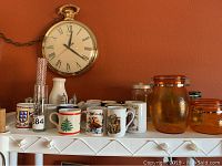 Overview of the lot showing coffee cups, mason-style amber jars with lids, small amber glass canisters, salt and pepper shakers, white ceramic vase and glass bottles on a white shelf against an orange wall with a large clock