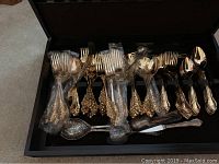 Photo of gold colored flatware set in a storage drawer, showing forks, spoons, knives with ornate detailed handles, some pieces wrapped in plastic