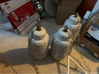 Three white ceramic Japanese hanging lanterns with lattice cut patterns, displayed on the floor with metal hanging chains extending out, surrounded by miscellaneous household items.