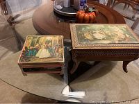 Two ornate vintage jewelry boxes on a glass table including a larger carved wooden box with a classical scene on top and a smaller box with a painted dance scene under glass