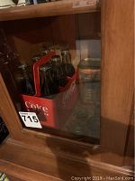Red Coca Cola plastic crate holding 6 glass Coke bottles with metal caps inside a glass display case
