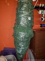 Green artificial Christmas tree tightly wrapped in clear plastic, upright view, with white wall and orange background.