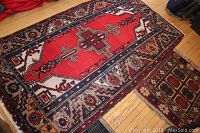 Photo showing the handwoven area rug with red central medallion and geometric border patterns on wood floor, with visible wear.