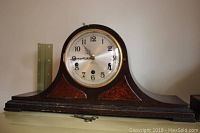 Front view of wood mantle clock with round face and Arabic numerals showing the time, sitting on a flat surface with winding key in front.
