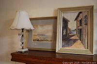 Table lamp with glass and brass base and two framed watercolour paintings on a wooden surface.