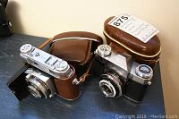 Two vintage 35mm cameras on a surface: Kodak Retina with brown leather case (closed) and Zeiss Contaflex with brown case behind, Kodak camera shows silver and black body with lens extended.