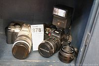 Photo showing the black Pentax Program Plus camera with mounted flash and black zoom lens next to the silver Pentax MZ-5n camera with 28-80mm lens and an additional lens in front.