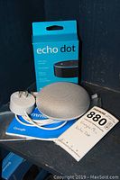 Google Home Mini smart speaker, power cord, and user booklet alongside Amazon Echo Dot in original blue box