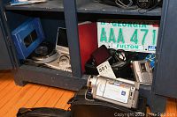 Photo of all items located on and under a dark shelf, shows digital camcorder, cassette recorder, CD player, GPS unit, car speakerphone, service manual, and vehicle license plate.