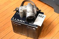 Silver Panasonic Lumix DMC FZ7 digital camera atop original box with MaxSold tag visible.