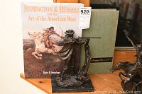 Photo of bronze sculpture of a man aiming a rifle in front of two books; one book titled "Remington & Russell and the Art of the American West" with illustrated cover showing Native American on horseback, and one large green hardcover book titled "Frederic Remington."