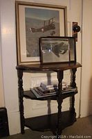 Photo showing the wooden demilune table with two lower shelves holding magazines and a framed aircraft print, along with a larger framed color biplane print mounted on wall.