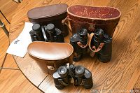 All three pairs of vintage binoculars with their cases displayed on a wooden table; varying case colors: brown and black leather.