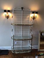 Front view of white metal and brass bakers rack showing two shelves, one with broken glass, decorative scrollwork and brass accents
