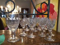 One large Waterford crystal glass with detailed cut patterns and 11 smaller crystal liqueur glasses lined up on a wooden surface.