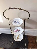 Full view of brass 3-tier stand holding three oval plates each with a shoe motif, standing on a dark floor mat against wall.