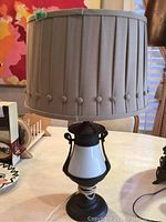 Full view of lamp showing dark metal and white ceramic base with pleated beige lampshade and power cord on table
