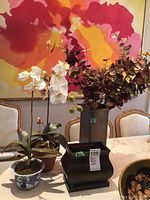 Wide view of artificial flower pots and metal vases on dining table, showing arrangement and scale.