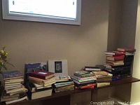 Wide shot of multiple stacks of books arranged on a wooden bench or low table showing varied titles and wear