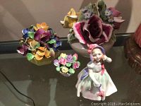 Overview of Royal Doulton figurine alongside coalport and china flower arrangements on a glass table.