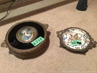 Two decorative plates on the floor, one round metal base with glass lid and one Asian style brass plate holder.