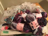 Five skeins of Patons Pirouette yarn in a variegated colorway on a table with cross stitch supplies in plastic bags.