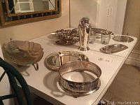 Collection of silver plated serving dishes, trays, coffee pot, glass condiment jars on a white table, showing condition and wear.