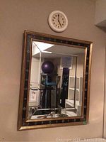 Photo of framed mirror on wall with black and gold trim, presence of a round battery operated wall clock above it