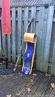 CCM hockey stick leaning against a wooden fence next to a Torpedo toboggan sled with blue fabric seating on a wooden deck with leaves scattered around.
