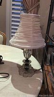 Clear pressed glass lamp base and white ruched fabric shade on a table near a window with beige curtains, showing the overall lamp design and condition.