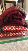 Folded large red hand-knit wool blanket with multicolor accents on top of two square red pillows with white circular linked chain pattern.