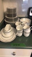 Stacked Noritake dinner plates, salad plates, dessert plates, soup bowls, cups and saucers arranged on countertop showing quantity and condition.