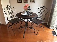 Photo shows the 3 piece bistro set with a round wooden table top, wrought iron base, and two matching wrought iron chairs with wooden seats.