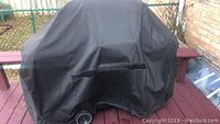 Outdoor propane gas BBQ grill fully covered with a black protective cover on a wooden deck.