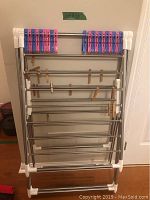 Full view of the foldable metal clothes drying rack with multiple horizontal bars and plastic clothespins clipped on the top bar, additional wooden clothespins on lower bars.