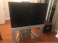 Front view of Samsung 42 inch DLP TV on silver metal stand with glass shelf, including power cable plugged in.