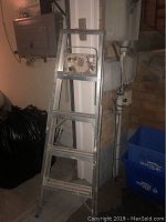 5 foot aluminum step ladder leaning against a wall in a basement area, showing the four wide steps and the top platform with some marks and paint spots.