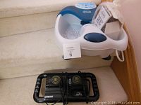 Photo showing Dr. Scholl Foot Bath Spa with remote control box and Conair handheld massager placed on carpeted stairs.