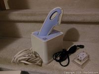 Hair removal device in charging base with power cord and adapter plug visible on carpeted stairs