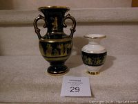 Two handmade Greek vases displayed on stairs. One is larger black with gold figures and handles, the other smaller white with black and gold Greek figure band.