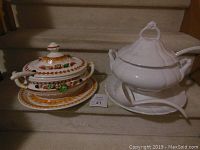 Two ceramic soup tureens with lids, matching under plates and ladles, placed on carpeted stairs; one oval chili/stew tureen decorated with painted vegetables and fruits, the other plain large white soup tureen.