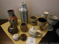 Full view of 11 handmade pottery pieces on a yellow cloth, including vases, cups, goblets, teapot, and shakers.