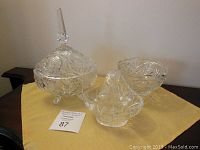 Three clear glass items on a yellow cloth: 7-inch oval candy dish with lid, 6-inch round bowl, and 6-inch basket with handle, all featuring intricate cut glass designs with pinwheel patterns.