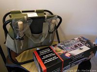 View of green garden caddy chair with black metal frame, beige accents, and five garden tools with wooden handles in plastic packaging clipped to the side pockets. Box of car wash system hose attachment placed beside chair.