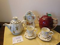 View of all items including white porcelain teapot and cups with gold detailing, floral decorated clock, and red apple-shaped ceramic teapot and cup.