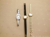 Three ladies watches displayed on fabric background; silver Wyda watch, gold Creation watch with black strap, gold Wittnauer watch with linked bracelet