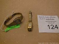 Two ladies wristwatches displayed on surface with auction lot card in background. One Bulova Accutron watch with gold-tone bracelet and one smaller Seiko watch.